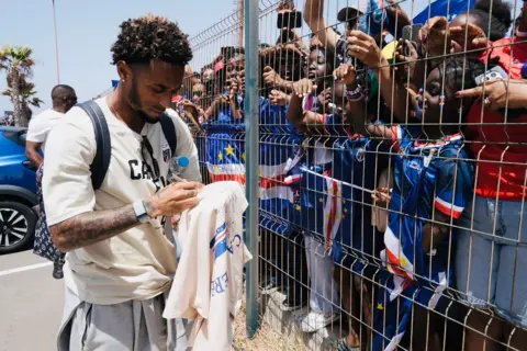 Thousands welcome home Cape Verde footballers after stunning World Cup run Michel Mvondo/BBC Captain Ryan Mendes signs a white shirt, while separated from fans by a metal fence