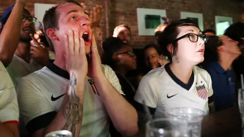 Getty Images Two United States football fans react during the match between Ghana and USA at the WoodWork bar in the Brooklyn borough of New York City. One is a man and the other is a woman. They are both wearing USA football tops.