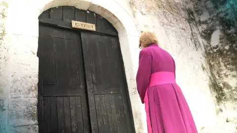 Dame Sarah Mullally from behind. She is wearing pink robes and hair short blonde hair. She is facing a large brown wooden door which has a small golden plaque on it with the words 'door of return' engraved.