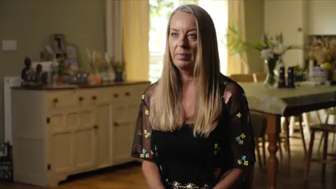 Sacha Wall, a woman with long blonde hair, wearing a black top with flowers on the arms, sitting in a large kitchen-diner, with a table and sideboard behind her.