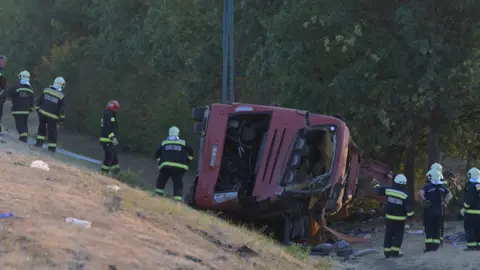 EPA A crumpled red bus sits at the bottom of a ravine as firefighters attend.
