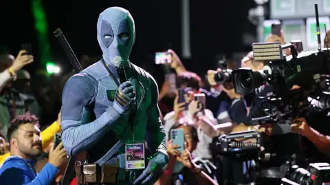 Getty Images Ryan Reynolds, disguised in a Deadpool costume, holds a microphone and is surrounded by cameras and fans as he speaks at San Diego Comic Con.