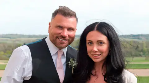 Dave Howell and BeccaGibson Smartly dressed man in waistcoat, shirt and pale pink tie, next to a woman with long hair against a backdrop of green countryside. Both are smiling