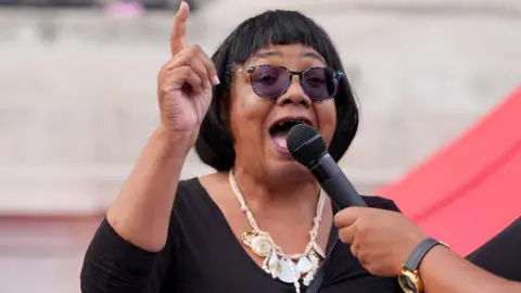 PA Media Labour MP Diane Abbott speaks into a microphone at a Trafalgar Square vigil staged to commemorate Jason Arday. She is dressed in black and wears dark glasses and white jewellery.