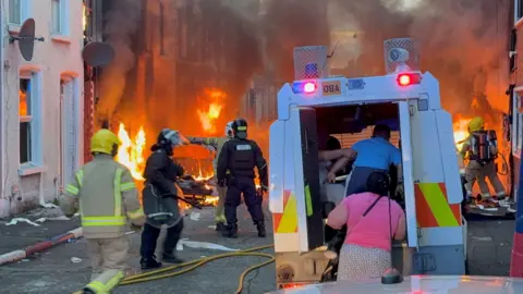 PA Images A riot-control police vehicle is parked in a narrow street as firefighters battle multiple fires around burning cars and buildings. Thick smoke fills the area while emergency crews and a few civilians move through the scene amid debris and fire hoses.