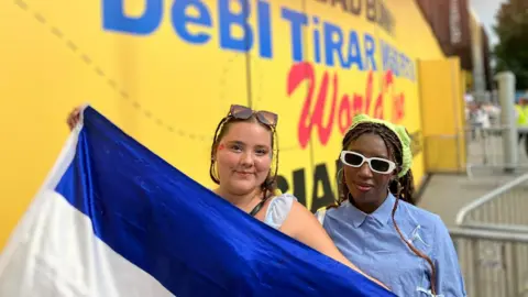 Two young women stand outside a stadium. They both have sunglasses on. The woman on the left is holding a blue and white Honduran flag, covering her outfit. The woman on the right is wearing a blue buttoned up shirt and a green bandanna.   
