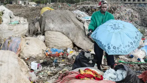East Africa wants to curb imports of used clothes, but it's not easy AFP via Getty Images A man in a green jacket holds a blue umbrella at a landfill site full of bails and piles of second-hand garment in Nairobi - October 2025.