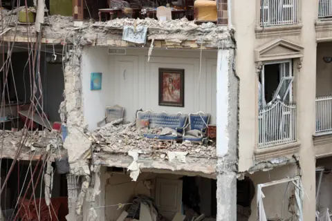 EPA/Shutterstock A partially destroyed residential apartment block in Tehran. The outside wall is no longer present and a blue sofa with gold trim can be seen in a room filled with rubble. There is a picture still hanging above it on the wall.   