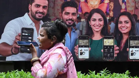 LightRocket via Getty Images A woman wearing a pink Indian dress speaks on phone near a poster of a mobile phone advertisement in Mumbai city, with four models showcasing their smartphones. 