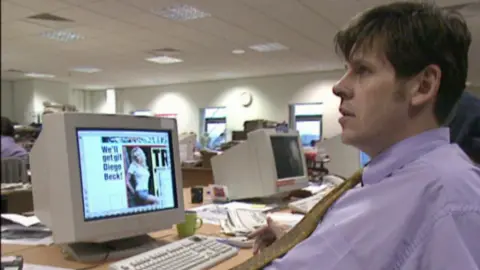 Tony Livesey, pictured in the 1990s for an episode of the BBC TV show Back to the Floor, with his hair cut in a fringe, in shirtsleeves and wearing a gold tie, sitting at a desk in front of a bulky computer monitor showing a desktop publishing programme with a page of one of the Sport newspapers displayed on it.