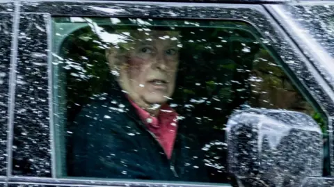 Bav Media Andrew Mountbatten-Windsor wearing a pink shirt and dark jacket pictured behind a rain-spattered window of his vehicle