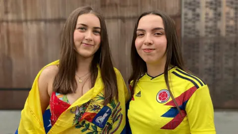 Two girls stand side by side. One is wrapped in a Venezuelan flag. The other is wearing a yellow Colombian football shirt. 