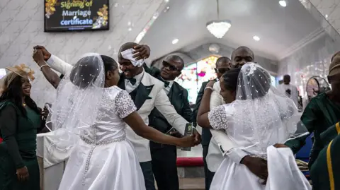 AFP via Getty Images The grooms and brides dance during their wedding ceremony. A man wearing sunglasses wipes one of the groom's brow with a white cloth.