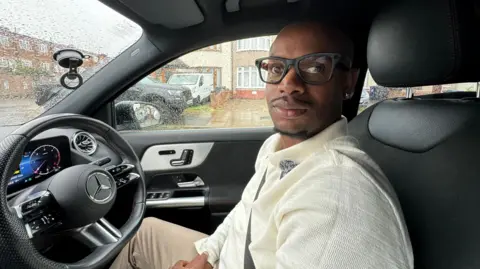 Donovan Smith, a driving instructor in south London sits at the sterring wheel of his car. He has shaved hair and wears glasses, wearing a cream summer jumper and beige trousers. 