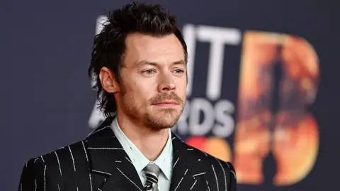 Getty Images Harry Styles wears a pinstripe suit with a light green shirt and silver tie underneath. He stands against a Brits backdrop