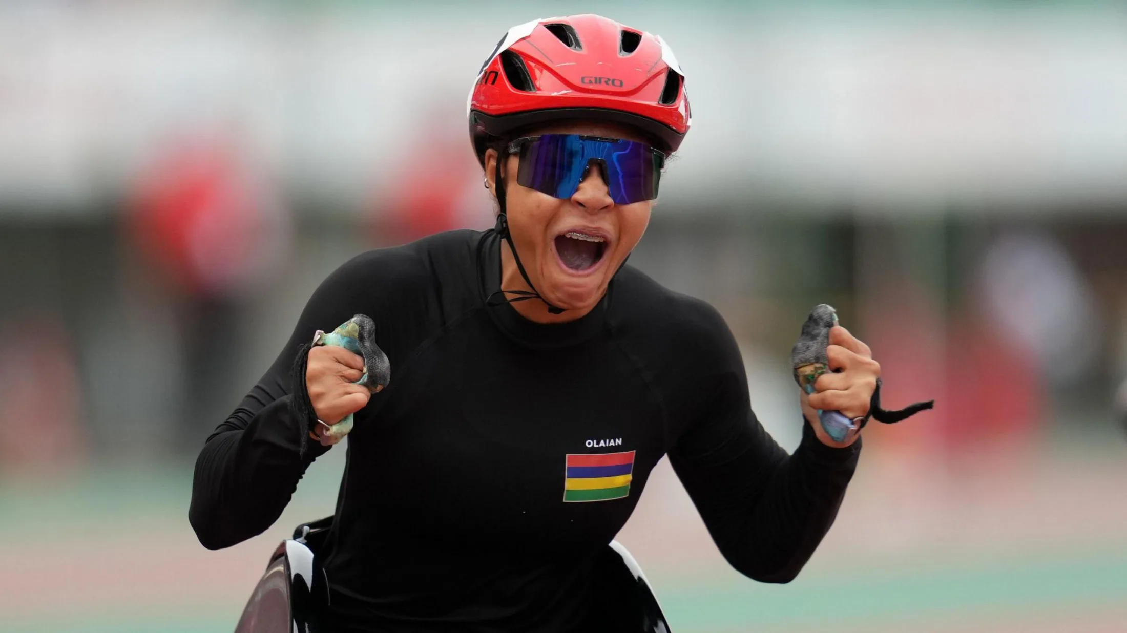 Noemie Alphonse clenches her fists and screams in delight as she finishes a wheelchair race. She wears a tight black long-sleeved sports top with the Mauritian flag on its left breast, big mirrored sunglasses and a red racing helmet. Just the very top of her wheelchair is peeking into view at the bottom of the image