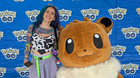 10 years of Pokémon Go and the millions still trying to catch 'em all j0beats A picture of the Pokémon GO content creator j0beats who has blue hair, and is smiling next to a large costumed Eevee Pokémon against a blue backdrop which says Pokémon Go. She is wearing a black and white cow style top and a denim skirt.