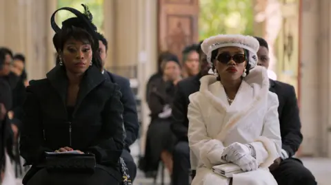 The Polygamist's creator says women see themselves reflected in her Netflix hit Netflix/ Iconic Digital Two women in elegant formal attire sit side by side, one in a black coat with a feathered fascinator hat and the other in a cream fur-trimmed coat with a matching hat, sunglasses, pearls, and white gloves. There are other seated guests in dark clothing behind them.
