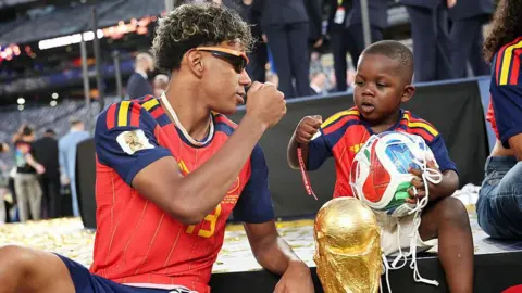 Hector Vivas - FIFA / Getty Images Yamal fist bumps his younger brother in front of the World Cup trophy