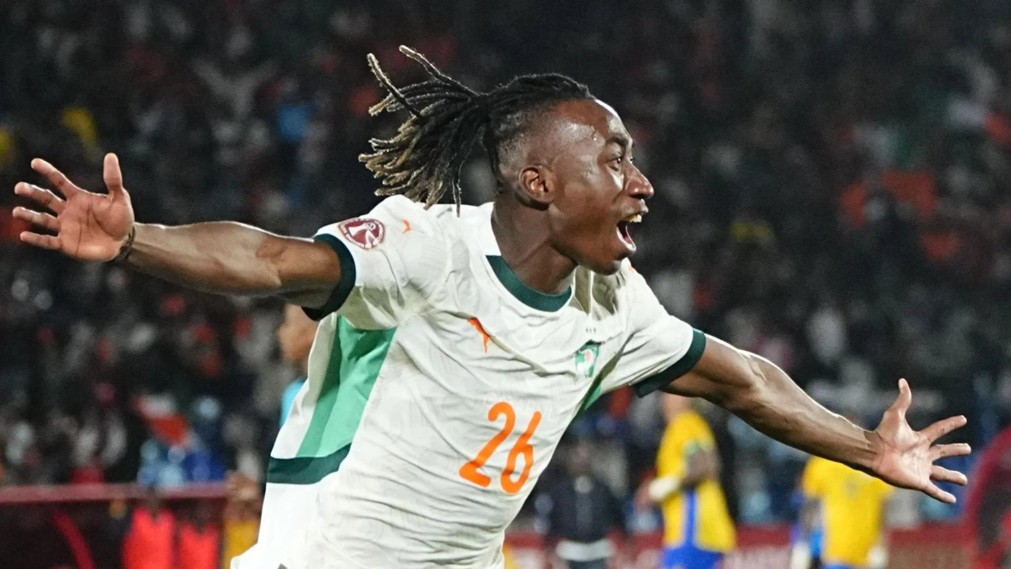 Yan Diomande, wearing a white Ivory Coast shirt with an orange number 26 on the chest, spreads his arms wide and opens his mouth as he wheels away in celebration, his hair seen flowing behind him. A packed crowd in a football stadium is out of focus in the background
