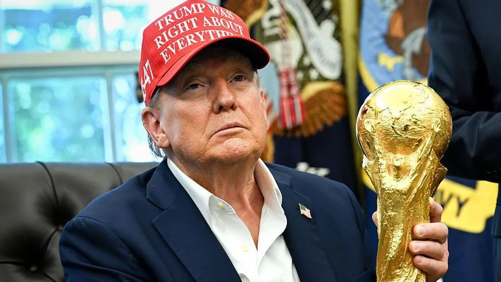 US President Donald Trump visible from chest up holds the Fifa World Cup trophy while sitting in a black chair in the Oval Office in the White House. He wears a blue suit jacket bearing an American flag lapel badge, an open-necked white shirt and a red baseball cap bearing the message 'Trump was right about everything'. Out of focus in the background are a window and two flags