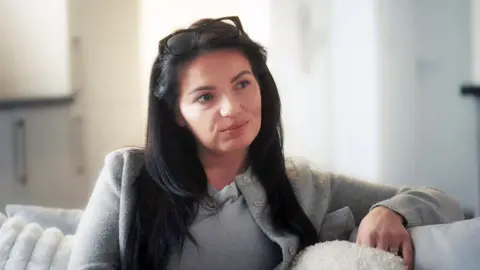 Leanne sits on a white sofa looking at someone off camera with a serious expression. She has long dark hair and glasses perched on the top of her head.