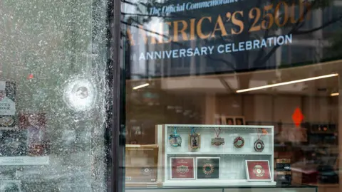 Getty Images A bullet hole can be seen in the fractured glass window of the White History shop after yesterday's shooting near the White House. Through an intact window pane, one can see a sign declaring, "America's 250th Anniversary Celebration."