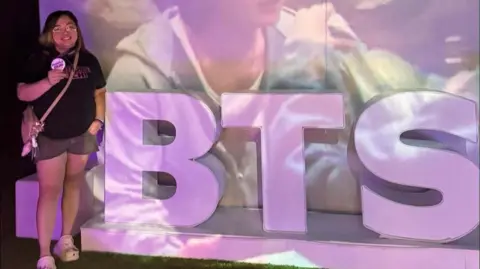 The BTS fans losing thousands as scammers cash in on comeback tour 'ticket war' Supplied Cookie poses beside a purple BTS signage at an event organised by Army