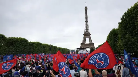 AFP via Getty Images GV image showing PSG fans waving flags with the club's logo on, with the Eiffel Tower in the background