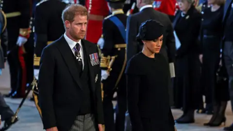 Getty Images Mid-shot of Prince Harry and Meghan Markle at the Queen's funeral in 2022. Prince Harry is wearing a black morning suit with a white shirt and black tie with military medals on his left side. Meghan is wearing all black.