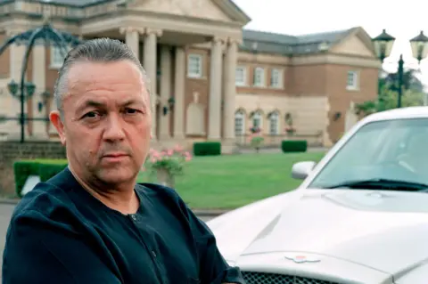 David Sullivan, wearing a black T-shirt, poses with his arms folded next to a silver Bentley outside his home, Birch Hall in Essex, a large neo-classical mansion with an imposing stone portico supported by pillars. He was pictured in 2003 for a BBC documentary called Sex Empires: Rise of the Porn Barons