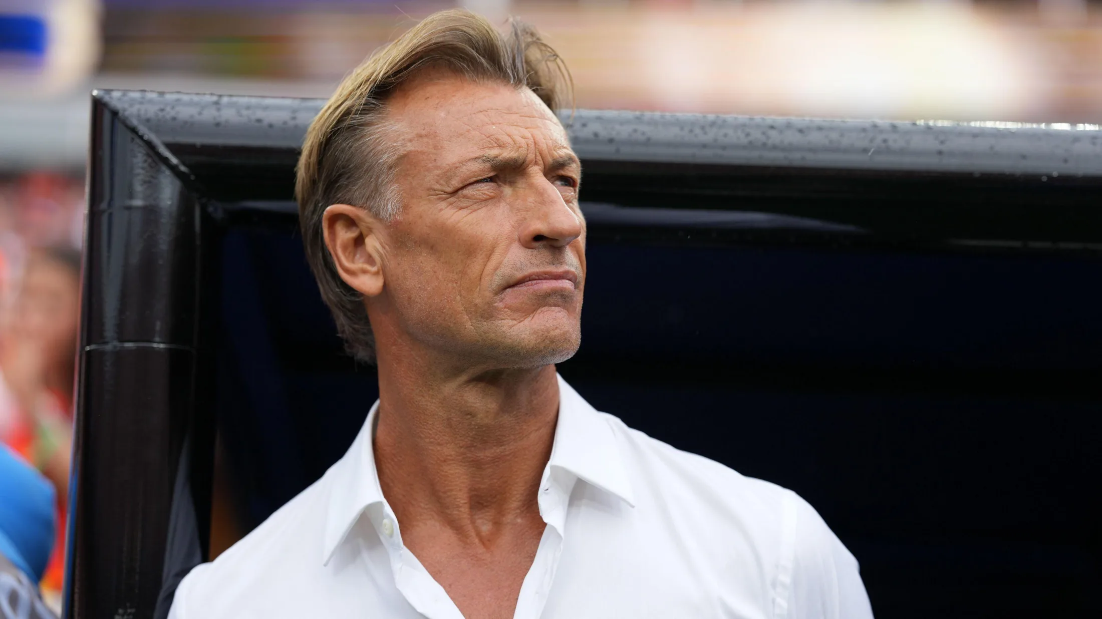 Herve Renard, visible from shoulders up while wearing a white shirt open at the neck, looks slightly to his left and holds his head high with his eyes staring into the distance as he stands in front of a black dugout at a football stadium