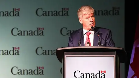 Getty Images An image of Tim Hodgson, Canada's minister of natural resources, speaking at a podium. Behind him is a backdrop with a repeated Canada logo, featuring the word Canada and a small Canadian flag above the letter 'a'. He is wearing a suit, and has combed over grey hair. 