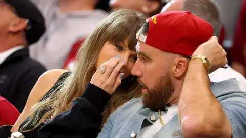 Gregory Shamus/Getty Images Close up of Taylor Swift whispering behind her hand to Travis Kelce who is wearing a red cap pointed backwards