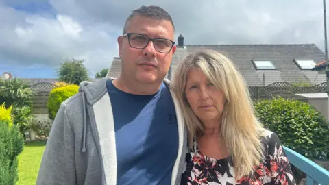 Brian Buckle ar y chwith. Mae ganddo wallt byr llwyd a'n gwisgo crys-t UnderArmour glas a hoodie llwyd gyda zip. Mae Elaine Buckle ar y dde, mae ganddi wallt hir melyn a chrys du â blodau coch a Gwyn. Mae'r ddau yn sefyll yn yr Ardd ac yn edrych mewn i'r camera.