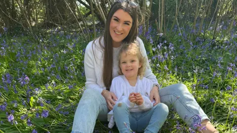 Other Alexandra is sitting outside on the ground surrounded by bluebells with three- year-old Ivy in front of her. They are both wearing white tops and pale blue jeans and are looking into the camera and smiling.