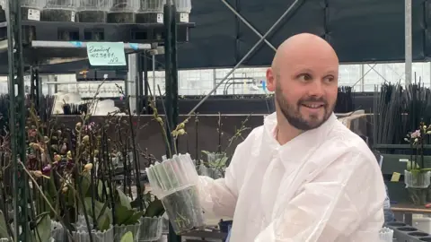 Stefan Kuiper wears a protective white suit and looks out of shot. Behind him are shelves of orchid plants. 