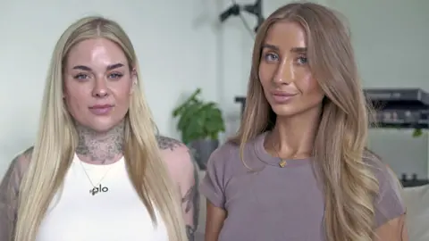 A head and shoulders portrait of OnlyFans creators Gia Clarke, on the left and Lily Phillips, on the right. They are looking into the camera smiling with their mouths closed. Gia wears a white tank top and has tattoos on her neck and shoulders, framed by long blonde hair. Lily has wavy light brown hair and a dark tshirt on.  
