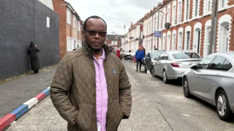 PA A man standing in a street lined with terraced houses. He is wearing a brown anorak and pink shirt and wearing dark-lensed glasses. He is balding, and has some hair towards the back of his head. On the right side there is a line of cars parked in front of the houses. On the left the kerbstones are painted red, white and blue. It is an overcast day. 
