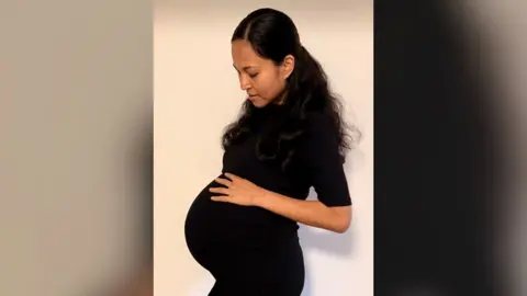 Jenitar Na’amoana/Gofundme Jenitar Na'amoana pictured while pregnant. She has long dark hair and is wearing a black short-sleeved dress. She is looking down and touching a very visible baby bump.