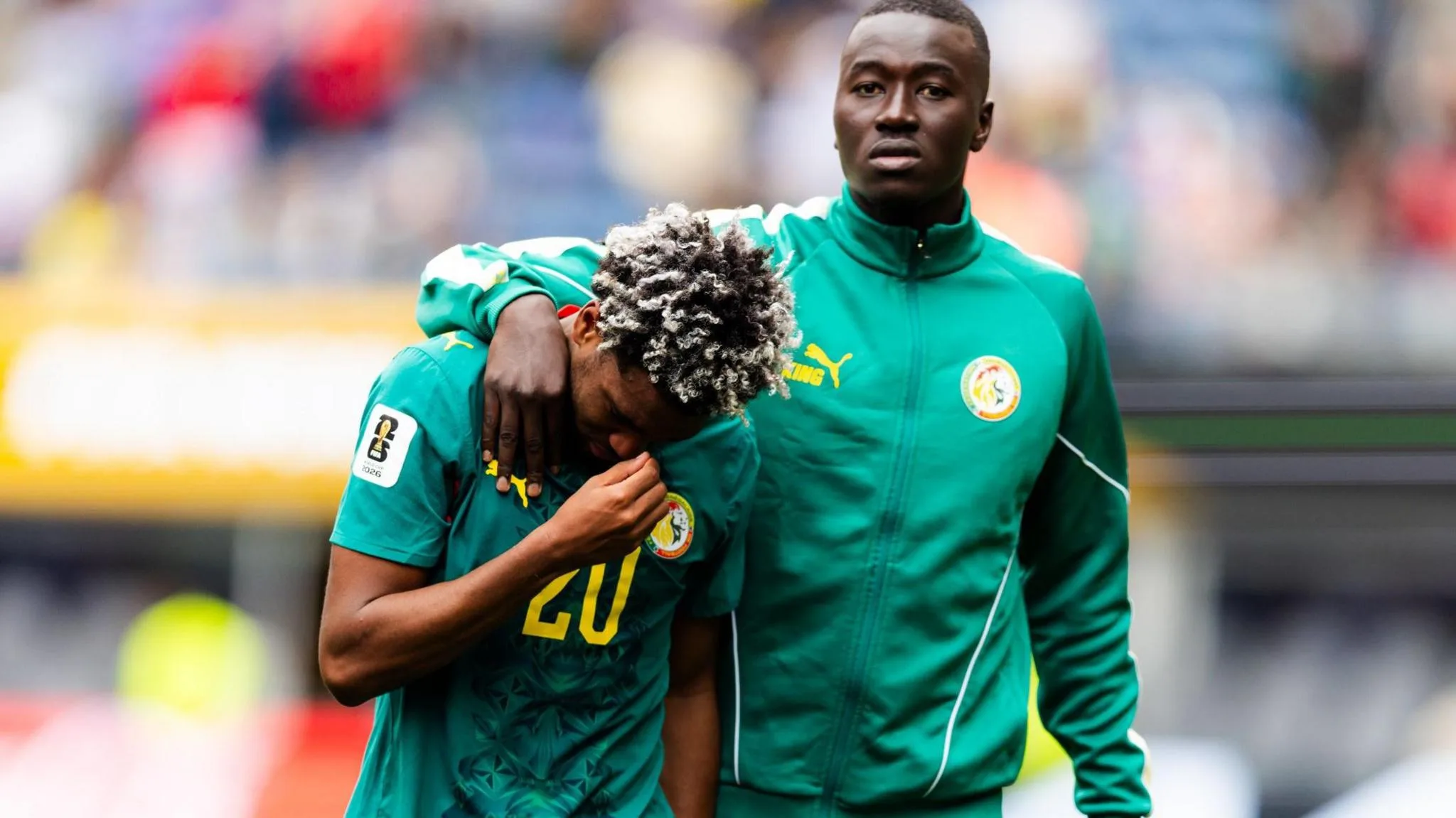 Pape Gueye, wearing a green Senegal tracksuit top, looks towards the camera as he consoles Ibrahim Mbaye with an arm around the shoulder of his team-mate who casts his face downwards and holds his right hand up to his face. Both players are visible from waist up and a stand in a football stadium can be seen out of focus in the background