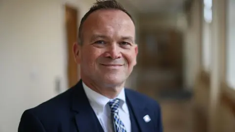 Secondary schools in Scotland cutting subjects due to teacher shortage 'crisis' Laurence Findlay, a man with short, brown hair, standing in a school corridor. He is wearing a blue suit, white shirt and patterned tie.