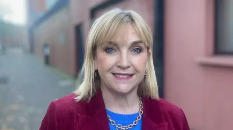 Why are so many organisations leaving X? Celine McStravick has shoulder-length blonde hair and blue eyes. She is smiling at the camera. She is wearing a blue jumper and a maroon blazer. Behind her is a red wall.