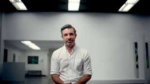 Brandon Silverman, a man with dark hair with a touch of grey, a side-parting and  stubble, wearing a white shirt and pictured sitting down in what appears to be a large gallery space with mostly bare white walls and bright overhead lighting.