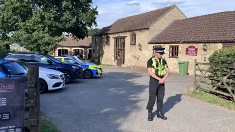 Sam Read/BBC A uniformed police officer stands in front of a bricked barn has a car park outside the front on the left. There is a fence on the right which opens into a court yard and there are signs on the door. There is a police car parked in the car park.