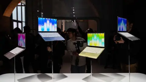 Bloomberg via Getty Images A set of colourful Apple Neo laptops positioned at varying heights on stands in a display at an Apple Store, with photographers crowding around to capture them images of them.