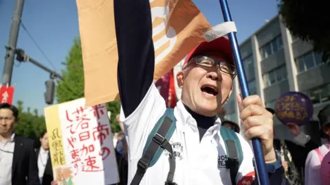 Anti-war protests rock Japan as PM pushes for stronger defence BBC News: Jiro Akiba A man in a backpack, glasses and a red cap punches a hand in the air while holding a flag