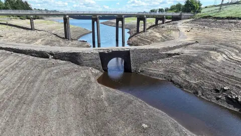 Getty Images Dried-up reservoir bed, with a small stream instead of a large expanse of water. A bridge is in the background and there is a light blue sky with scattered clouds.