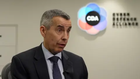 Dozens of romance scammers arrested after specialist fraud squad investigation in UK, Nigeria The head and shoulders of a man with short grey hair, wearing a blue jacket, shirt and tie, with a CDA logo blurred in the background.