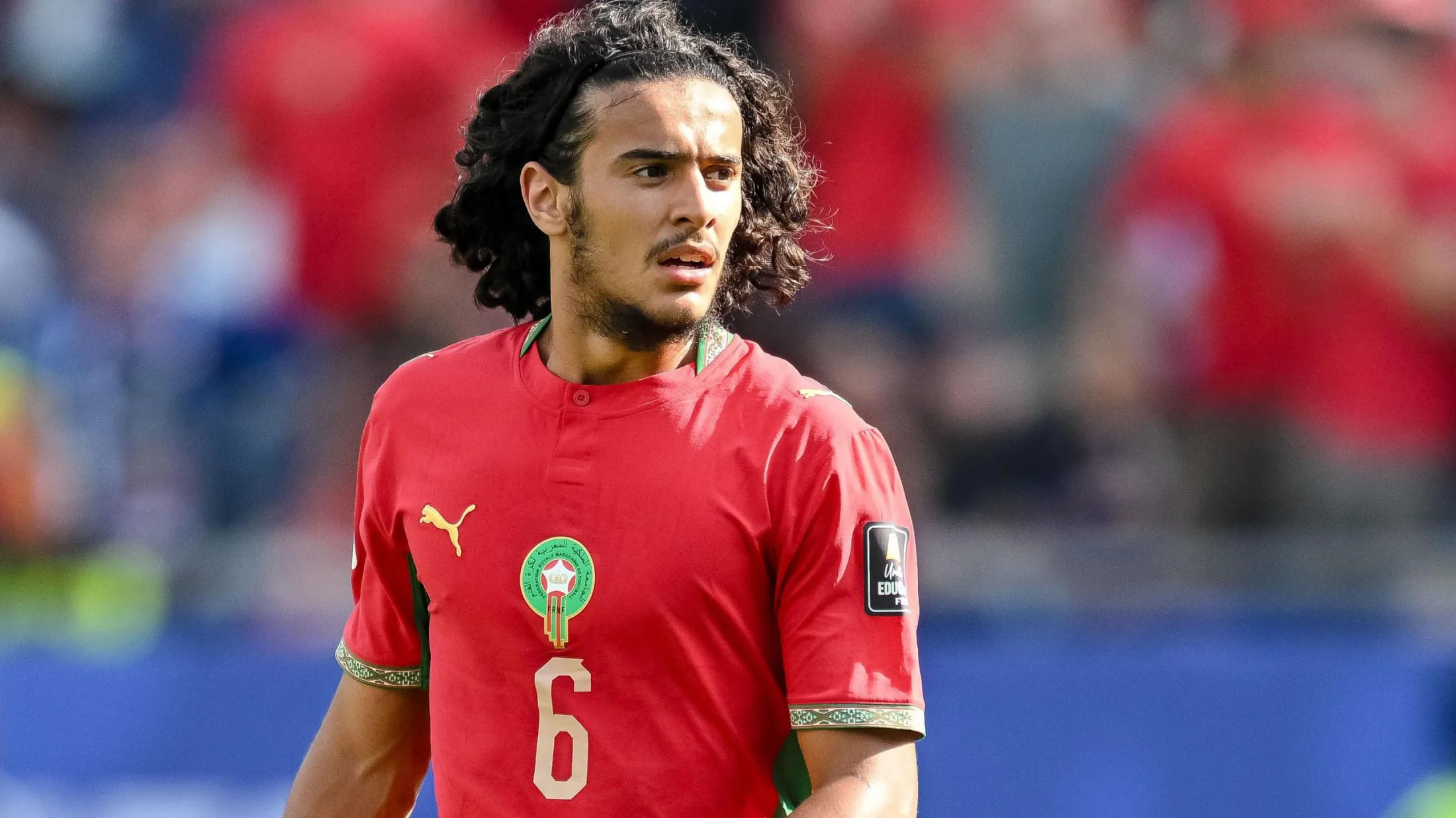 Ayoub Bouaddi, visible from waist up wearing a red Morocco shirt with a gold number six and federation crest on his chest, looks to his left during a football match. A stand accommodating fans can been seen out of focus in the distance